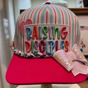 Può includere: Un cappellino trucker con visiera rosa e corona a righe verticali azzurre e bianche. Una toppa mostra la scritta RAISING DISCIPLES in lettere ricamate colorate con un cuore rosa. Sono presenti un bordo intrecciato e un fiocco con paillettes.