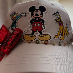 May include: White baseball cap with embroidered Mickey Mouse, Donald Duck, and Pluto designs. The cap includes a red sequined bow and a floral trim. A playful accessory for Disney enthusiasts.