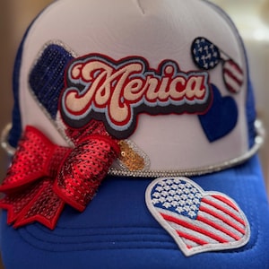 May include: A patriotic trucker hat with a white front panel and a blue mesh back. The hat features the word "Merica" in a retro font, a sequined red bow, and patches of the American flag and hearts.