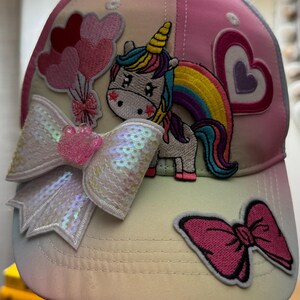 May include: A pastel-colored baseball cap with a unicorn and rainbow design. The cap features a sequined bow, heart and balloon patches. The unicorn has a yellow horn and a rainbow mane and tail. The cap is a mix of pink, white, and yellow.
