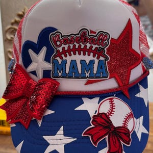 May include: A baseball cap with a white mesh back and a blue and white star-patterned front. The cap features a red sequined bow, a baseball graphic, and the text "Baseball MAMA" in a patch with red and blue accents.