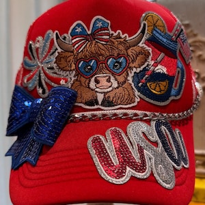 May include: A bright red trucker hat featuring a central embroidered patch of a brown highland cow wearing heart-shaped glasses and a patriotic bow. The cap includes a large blue sequined bow, a silver chain trim, and red and silver sequined text.