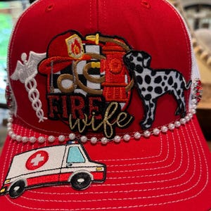 May include: Red and white trucker hat with a white pearl trim. The hat features embroidered patches of a firefighter's helmet, a fire hydrant, a dalmatian dog, an ambulance, and the words "Fire Wife".