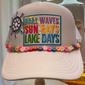 May include: A white trucker hat featuring an embroidered patch with the text BOAT WAVES SUN RAYS LAKE DAYS in colorful letters. To the left is a navy blue ship wheel patch. A string of small, multicolored clay flower beads lines the brim.