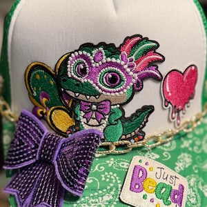 Puede incluir: Gorra de camionero blanca y verde con ala verde con estampado de cachemira. La gorra presenta un dragón de dibujos animados con una máscara morada, un lazo morado, un corazón rosa goteando y el texto "Just Bead It".