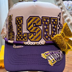 Könnte beinhalten: Eine violett-weiße Trucker-Cap mit den Buchstaben LSU in gelben und violetten Mustern. Die Kappe ist mit einer Tigergrafik, einer goldenen Pailletten-Schleife und einer violetten Perlenborte mit dem Schriftzug GAME DAY versehen.