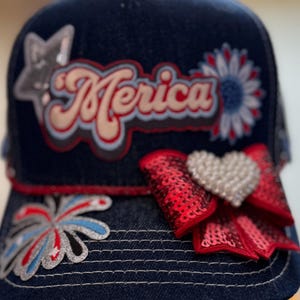 May include: A dark blue denim baseball cap with patriotic decorations. The cap has the word "Merica" in a retro font, a sequined red bow with a heart, and star and firework patches.