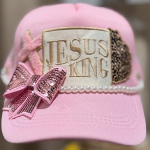 Può includere: Cappellino da baseball rosa con fiocco di paillettes, bordatura di perle e toppa con la scritta dorata "JESUS KING". Il cappellino presenta anche un dettaglio con stampa leopardata e un ornamento a forma di stella.