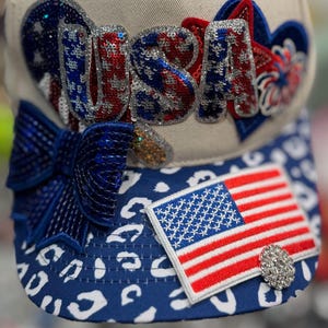 May include: A baseball cap with a white crown and a blue and white patterned brim. The cap features the word "USA" in red, white, and blue sequins, a small American flag patch, and a jeweled embellishment.