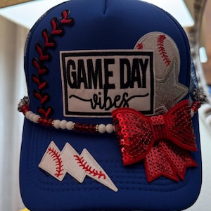 May include: Blue baseball cap with "GAME DAY vibes" text, baseball and star patches, and a red sequin bow. Features red stitching and a beaded band. A stylish accessory for sports fans.