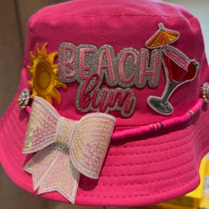 Pode incluir: Um chapéu de balde rosa brilhante com o texto "Beach Bum" em glitter prateado. O chapéu apresenta um laço de lantejoulas, um sol amarelo e um gráfico de cocktail. Um acessório divertido para a praia ou piscina.