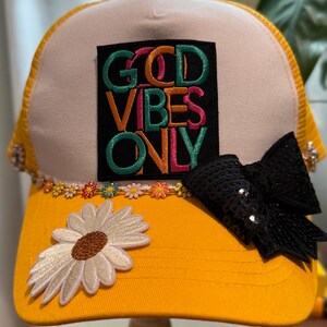 May include: A yellow and white trucker hat featuring a black rectangular patch with the text GOOD VIBES ONLY in colorful embroidered letters. The hat is decorated with a row of small flower charms, a large white daisy applique, and a black sequined bow.