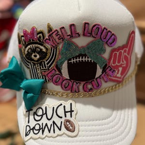 May include: White trucker hat with a football theme. Features a raccoon, a football, and the phrases "YELL LOUD LOOK CUTE" and "TOUCH DOWN". Embellished with sequins, a teal bow, and a gold chain.