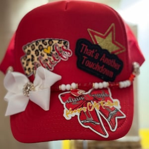 Puede incluir: Gorra de béisbol roja adornada con parches y adornos. Cuenta con un lazo blanco con un centro de pedrería, un acento de cuentas y parches con texto y un diseño de estampado de leopardo. El texto en los parches dice "Chiefs" y "That's Another Touchdown".