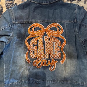 May include: A blue denim jacket with an orange sequined "GAME DAY" design on the back. The design features a bow and a checkered pattern within the letters. The jacket has a classic collar and button closures.