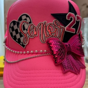 May include: A bright neon pink trucker hat featuring a black and white checkered heart patch with the word Senior in cursive script, a black star patch with the number 27, a large fuchsia sequin bow, a string of white pearls, and a silver chain.