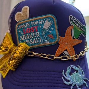 May include: A purple trucker hat featuring a gold sequin bow, a gold chain trim, and embroidered patches including a heart, a blue rectangle with the text LOOKIN FOR MY LOST SHAKER OF SALT, an orange starfish, a green margarita, and a blue crab.