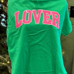 Puede incluir: Una camiseta verde brillante con la palabra "LOVER" en letras grandes, rosas y con lentejuelas en el pecho. La camiseta tiene mangas cortas y cuello redondo. La tela parece suave y cómoda.