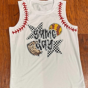 May include: White sleeveless shirt with a red and white baseball stitch design on the straps. The shirt has a black and white checkered star with the words "game day" in silver glitter letters and a red and yellow softball.