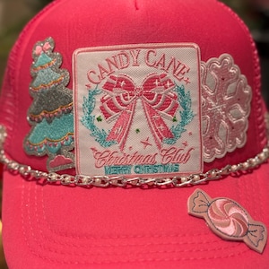Op de afbeelding: Felroze truckerpet met een mesh achterkant en een gebogen klep. De pet heeft geborduurde patches van een kerstboom, sneeuwvlok en zuurstok. De belangrijkste patch leest "Candy Cane Christmas Club" met een strik ontwerp. Een ketting met parels accentueert de klep.