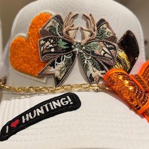 May include: A white baseball cap featuring an orange fuzzy heart patch, a camouflage bow with deer antlers, a gold chain link accent, a bright orange sequined bow, and a black patch with the white text I heart HUNTING!.