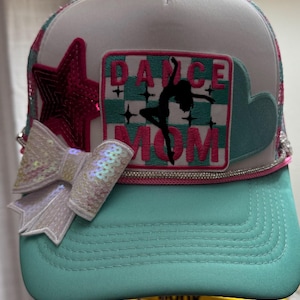 Puede incluir: Gorra de camionero turquesa y blanca con lazo de lentejuelas y estrella rosa. La gorra presenta un parche con las palabras "DANCE MOM" y una silueta de bailarina. Un ribete de pedrería adorna el borde.