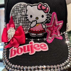 Könnte beinhalten: Schwarze Baseballkappe mit einem Hello Kitty-Aufnäher, einem pinkfarbenen Paillettenstern und einer pinkfarbenen Schleife. Der Rand ist mit Strasssteinen besetzt, und das Wort "boujee" ist auf der Vorderseite gestickt.