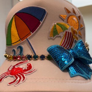 May include: Cream-coloured baseball cap adorned with beach-themed patches. Includes a rainbow umbrella, sun, crab, and a blue sequined bow. A jewelled band decorates the brim, adding a touch of sparkle to this summer accessory.