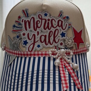 May include: A beige baseball cap with a blue and white striped brim. The front features a glittery red and silver patch with the text 'Merica y'all', accented by red and silver stars, a red gingham ribbon, and a cluster of white faux pearls.
