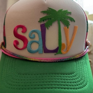 May include: A white foam trucker cap with a green peak and a rainbow-coloured cord trim. The front features the word "salty" embroidered in colourful letters, with a green palm tree replacing the letter "l".