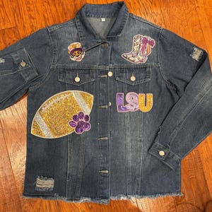 May include: A distressed denim jacket with multiple patches. Patches include a football, paw print, and the letters "LSU" in purple and gold. The jacket has a button-up front, two chest pockets, and a frayed hem.