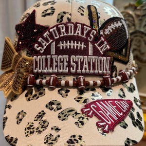 May include: A beige baseball cap with a leopard print pattern, featuring a maroon patch that reads SATURDAYS IN COLLEGE STATION with a football graphic. It includes a gold sequin bow, a maroon star, a football patch, and a GAME DAY pennant patch.