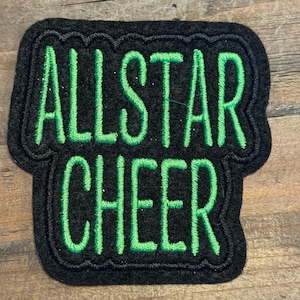 May include: A black fabric patch with a scalloped edge featuring the text ALLSTAR CHEER embroidered in bright green thread. The patch has a rectangular top section and a wider rectangular bottom section, creating a distinct shape.