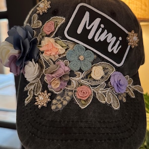 May include: A dark grey baseball cap decorated with fabric flowers in shades of blue, pink, and purple. The cap has the name "Mimi" in white lettering on a black patch, and is embellished with pearl accents.