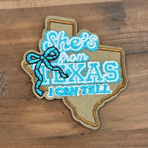 Könnte beinhalten: Ein beigefarbener, bestickter Aufnäher in Form des Bundesstaates Texas mit dunkelbraunem Rand. Der Aufnäher zeigt den hellblauen gestickten Text "She's from TEXAS I CAN TELL" und eine leuchtend blaue gestickte Schleife auf der linken Seite.