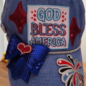 Könnte beinhalten: Eine Jeans-Baseballkappe mit patriotischen Verzierungen. Mit einer paillettenbesetzten blauen Schleife mit Herz, roten Sternen und einem Aufnäher mit der Aufschrift "GOD BLESS AMERICA" in Blau und Rot. Eine Strasskette akzentuiert den Rand.