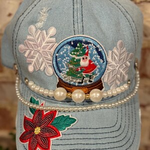 Op de afbeelding: Een lichtblauwe denim baseballcap met een parelrand en verschillende geborduurde patches. De patches omvatten een sneeuwvlok, een sneeuwbol met een kerstman erin en een kerstster.
