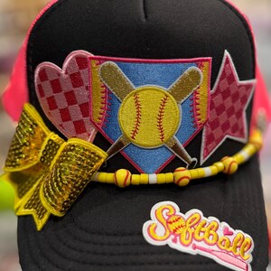 May include: Black and pink softball cap with a yellow bow, a beaded accent, and patches. The patches include a heart, star, and a shield with crossed bats and a softball. The word "Softball" is embroidered on the front.