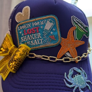 May include: A navy blue trucker hat with various embroidered patches, including a gold heart, starfish, crab, and a patch that reads "Lookin' for my Lost Shaker of Salt." A gold sequined bow and chain add embellishment.