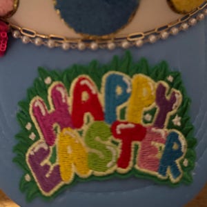 Könnte beinhalten: Eine blaue Baseballkappe mit weißem Schirm und einem bunten gestickten Aufnäher mit der Aufschrift "Frohe Ostern" auf grünem Gras.