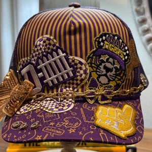 Gouden en paarse truckerpet in voetbalstijl. Dames. Tigers. Gouden schakelketting. Strass-steentjes, paillettenstrik en diverse patches. Thema: Defensie.