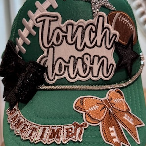 May include: A green baseball cap featuring the text "Touch down" in white and brown, a silver sequined star, a brown football patch, a black sequined bow, a brown bow with football stitching, and a banner reading "GAMETIME" in glittery letters.