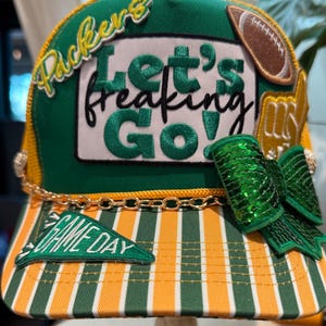 May include: A green and yellow striped trucker hat featuring a white patch with the embroidered text Let's freaking Go!, a Packers logo, a football patch, a gold foam finger, a green sequined bow, and a gold chain trim across the front brim.