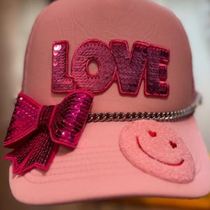 May include: Pink baseball cap with the word "LOVE" spelled out in sequined letters. The cap features a sequined bow, a silver chain detail, and a smiley face patch with heart eyes. A fun and fashionable accessory.