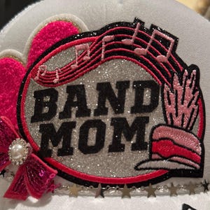 Puede incluir: Gorra de béisbol blanca con un parche de purpurina rosa y plata que dice "BAND MOM" con notas musicales y una pluma rosa y blanca.