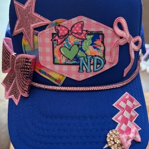 Puede incluir: Gorra de camionero azul con estrella rosa, lazos y un parche con estampado de cuadros rosas, diseño floral y las letras "ND". La gorra tiene una banda con pedrería y una joya decorativa.