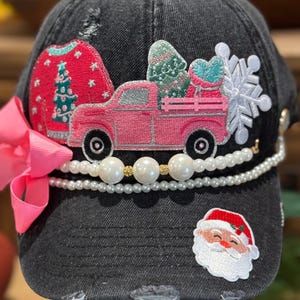 Puede incluir: Gorra de béisbol negra desgastada con adornos navideños. Cuenta con un lazo rosa, un parche de suéter rojo, una camioneta rosa que transporta un árbol de Navidad, un copo de nieve, un parche de Papá Noel y un ribete de perlas y cuentas doradas.