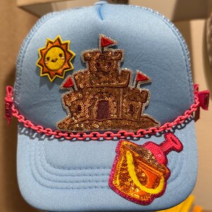 May include: Light blue trucker hat with a sandcastle, sun, and sand pail design. The sandcastle and pail are embellished with sequins. A pink chain detail accents the brim.