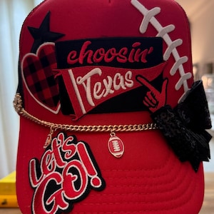 May include: A red trucker hat featuring a black patch with the text choosin' Texas, a red and black buffalo plaid heart, a white football lace graphic, a black sequined bow, a gold chain with football charms, and a patch reading let's go.