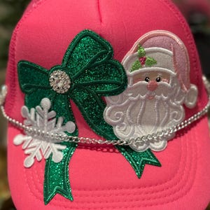 May include: Pink trucker hat with a green glitter bow, a Santa Claus patch, and a white snowflake. The hat has a silver chain around the brim. Festive Christmas-themed accessory.